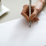 Close-up of female hand writing on blank piece of paper with cup of coffee in background