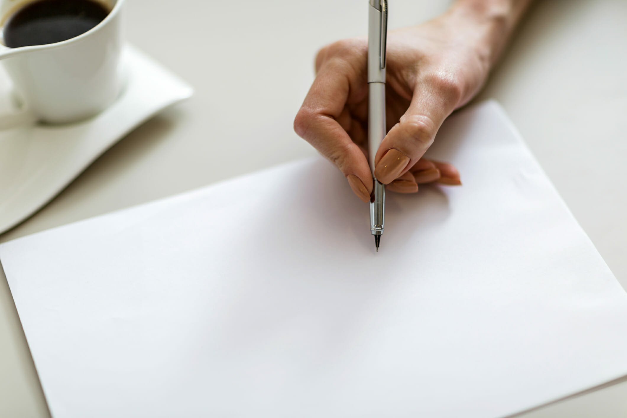 Close-up of female hand writing on blank piece of paper with cup of coffee in background