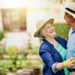 Senior man and woman smiling and dancing in their backyard while wearing sunhats and gardening gloves