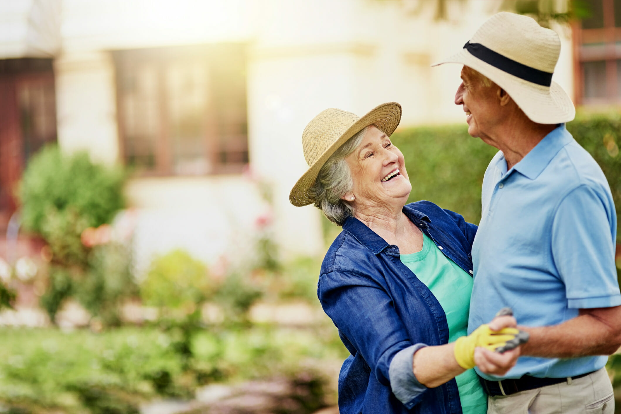 Senior man and woman smiling and dancing in their backyard while wearing sunhats and gardening gloves