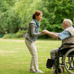 woman helps aging father stand out of wheelchair in park