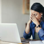 working woman at desk with laptop holding nose and looking like she has burnout