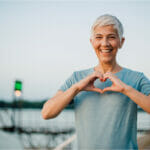 A mature lady doing a heart with her hand near a body of water