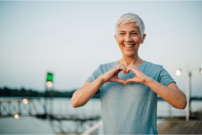 A mature lady doing a heart with her hand near a body of water