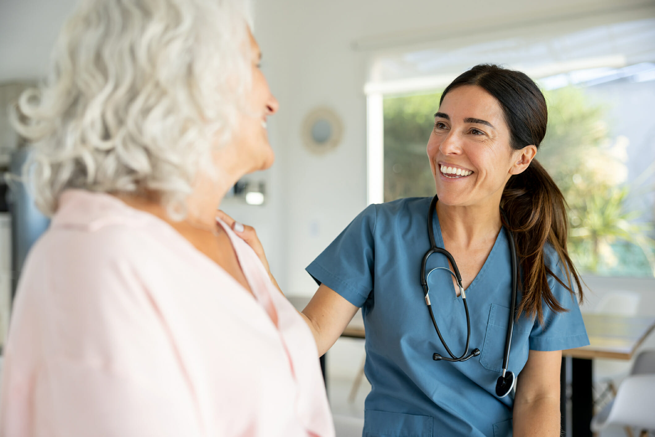 Happy nurse in blue scrubs with stethoscope talking to a senior woman on a house call and smiling